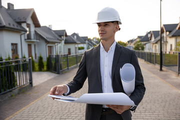 Real estate agent wearing hard hat and holding blueprints while standing in front of modern houses