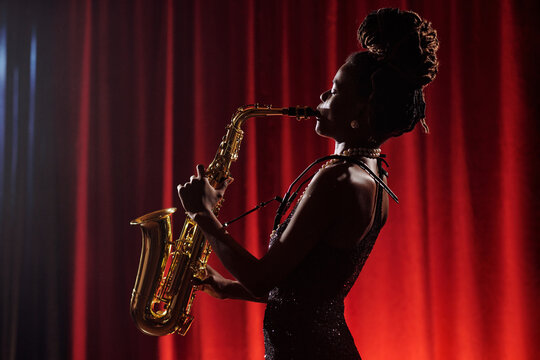 African American woman playing saxophone on stage in front of red curtain, standing in profile with hair in dreadlocks, performing jazz music as professional musician