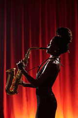 Black woman playing saxophone on stage in front of red curtain holding instrument with both hands...