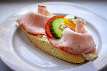 Open-faced sandwich with sliced ham, cucumber, red pepper, cheese, and creamy spread on baguette, served on a white plate.