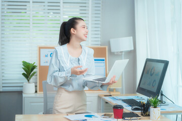 Confident beauty Asian office woman in a formal blouse hard working at her desk, reviewing a document with focus and a pen in hand. The workspace includes charts, a laptop, and a desktop computer.