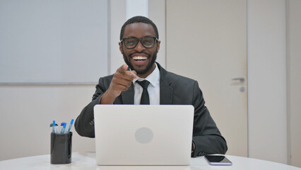 Engaged African American businessman in a suit, pointing directly at the viewer with an open mouth and wide-eyed expression in an office setting