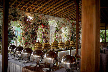 This image shows an elegant wedding buffet setup under a traditional wooden pavilion. The buffet tables are lined with polished silver chafing dishes and ornate golden food warmers, ready to serve a f