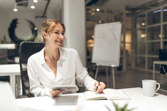 Businesswoman taking notes in notebook with whiteboard visible in office setting