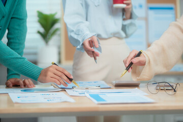 Three Asian women collaborate at shared office desk filled with documents, charts, coffee. One...