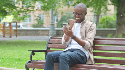 Obraz premium Young African Man Expressing Excitement While Using Phone on a Park Bench