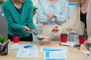 Three Asian women collaborate at shared office desk filled with documents, charts, coffee. One...
