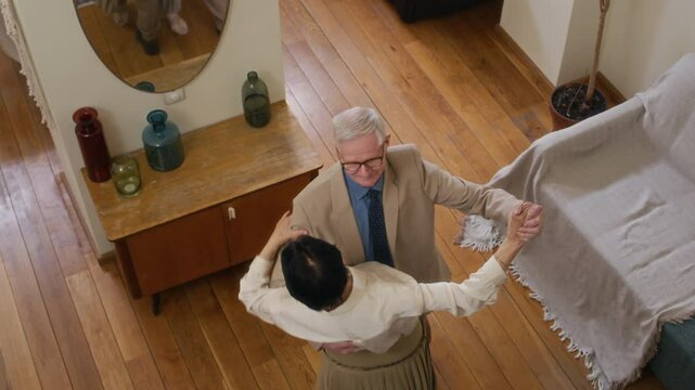 Slow motion of romantic elderly spouses wearing beige formalwear whirling in dance on wooden floor of living room with couch, mirror on wall and TV stand