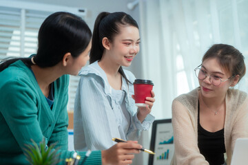 Three Asian women collaborate at shared office desk filled with documents, charts, coffee. One...