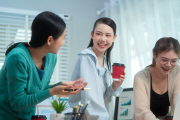 Three Asian women collaborate at shared office desk filled with documents, charts, coffee. One...