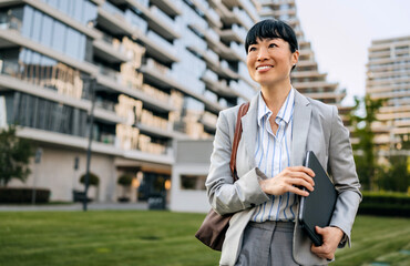 Professional Asian woman smiling while holding a tablet in a modern urban setting during the day