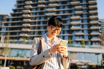 Smiling Asian woman engaged with smartphone in urban setting during sunny day