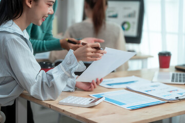 Three Asian women collaborate at shared office desk filled with documents, charts, coffee. One...
