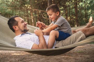 Father and son 5 years old fooling around playing relaxing in a hammock outdoors in a pine forest at a campsite. Tourism, active recreation, time spent with children, dad and son wrestle play