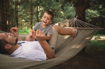 Father and son 5 years old fooling around playing relaxing in a hammock outdoors in a pine forest at a campsite. Tourism, active recreation, time spent with children, dad and son wrestle play