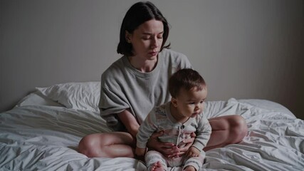 Tired young mother sitting on white bed, holding sleeping infant and experiencing emotional challenges of early parenthood, revealing raw moments of maternal struggle