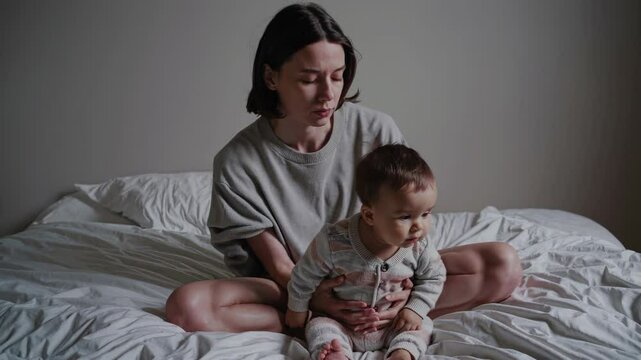 Tired young mother sitting on white bed, holding sleeping infant and experiencing emotional challenges of early parenthood, revealing raw moments of maternal struggle - Powered by Adobe