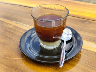 A cup of hot sweetened condensed milk coffee served in a glass on a black plate with a small spoon beside it, with a wooden table background, natural blur outdoor scene background.