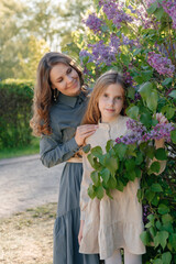 Fototapeta premium A happy mother and daughter in front of a blooming lilac tree. A family vacation on a sunny, warm day. Raising children, a happy family, and family values.
