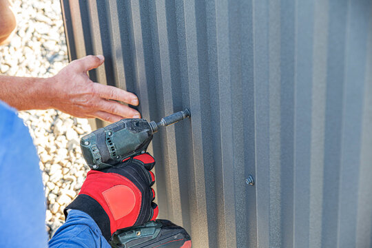A gloved construction worker uses a drill driver to drive a screw into a dark corrugated metal sheet to be installed on a fence or wall.