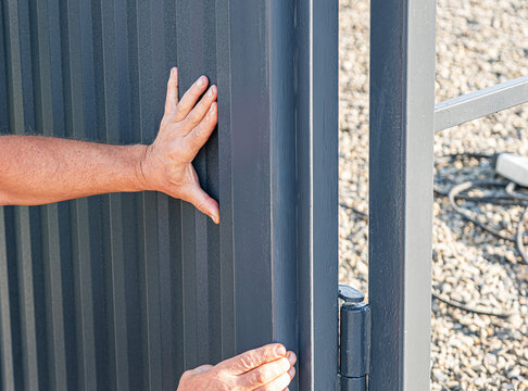 A person's hands are shown touching and adjusting a dark grey corrugated metal fence or gate panel next to a metal frame and hinge, with gravel on the ground.