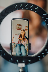 Young businesswoman recording video with ring light and showing clipboard