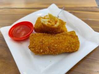 Crispy golden rissoles with savory and delicious filling, served on an elegant white plate, accompanied by a small bowl filled with bright red chili sauce and a fork, on the menu at an outdoor cafe.