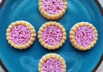 Round baked sweet biscuits with bright red filling and pink pastry sprinkles, on a decorative plate