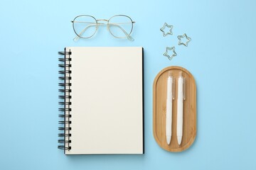 Notebook, glasses, pens and paper clips on light blue background, flat lay