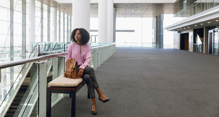 African American woman sitting on padded bench by escalator at expo with tan backpack, copy space