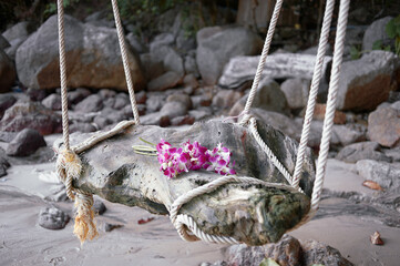A rustic wooden swing with purple flowers on a tropical beach, surrounded by rocks and trees.
