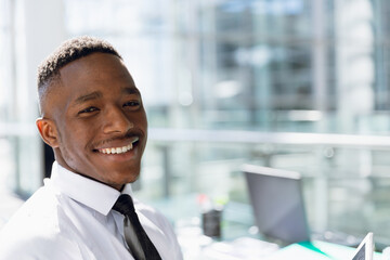 Smiling African American man wearing shirt and tie at office desk by window with laptop, documents