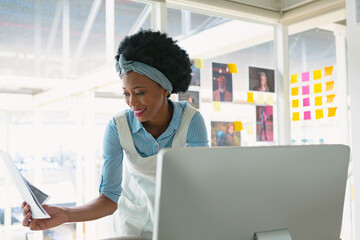 African American woman holding tablet and leaning over monitor in office with sticky notes, photos
