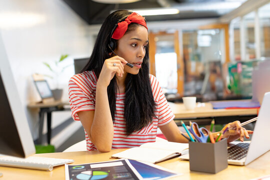 African American woman typing on laptop at office desk wearing headset and reviewing printed charts