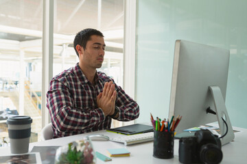 Male meditating with eyes closed at desk in modern office with computer, tablet, camera, pencils
