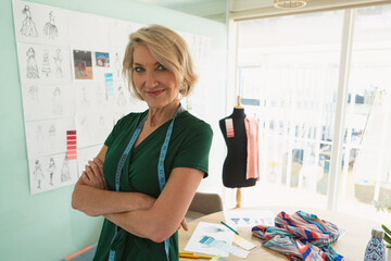 Mature woman wearing green wrap dress holding measuring tape posing in fashion studio with sketches