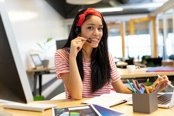 African American woman working in open office wearing headset and writing in notebook beside laptop