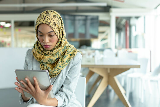 Middle Eastern woman wearing hijab and blazer using tablet at table in coworking office, copy space - Powered by Adobe