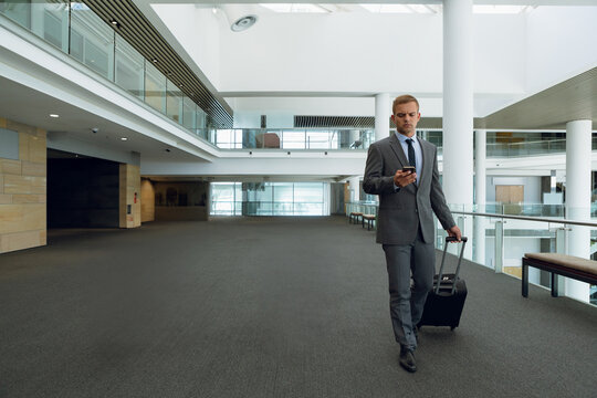 Male traveler walking through terminal using smartphone, pulling suitcase at railing, copy space