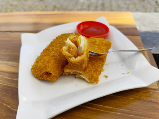 Crispy golden rissoles with savory and delicious filling, served on an elegant white plate, accompanied by a small bowl filled with bright red chili sauce and a fork, on the menu at an outdoor cafe.