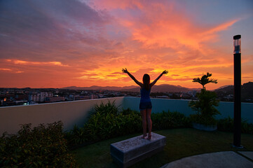 Fototapeta premium A person stands on a rooftop with arms raised, silhouetted against a vibrant sunset sky. The cityscape and distant mountains are visible in the background, creating a serene and uplifting scene.