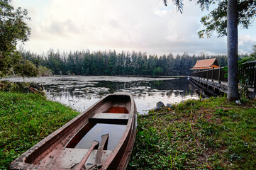 A serene lakeside scene at sunset with a wooden boat on the shore, a long pier leading to a house...