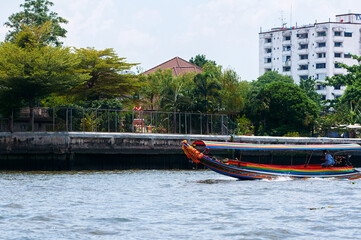 Obraz premium Colorful Longtail Boat Gliding by Riverside Urban Scenery Under Clear Sky