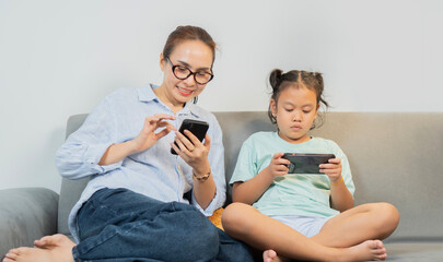 Little daughter and a mother sitting on sofa with a tablet and phone for online games at home