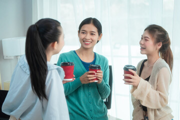 Three Asian women collaborate at shared office desk filled with documents, charts, coffee. One...
