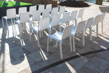 Rows of modern white plastic chairs are arranged outdoors on a sunlit stone patio, casting strong shadows. Ideal for concepts of events, gatherings, or minimalist design settings