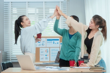 Three Asian women collaborate at shared office desk filled with documents, charts, coffee. One...
