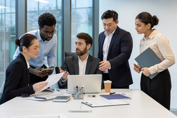 A diverse team of business professionals collaborating on a project in a modern office setting, using a laptop and documents.