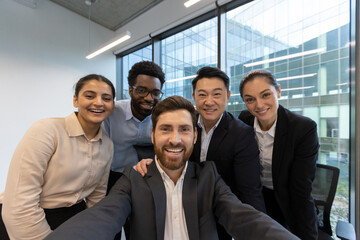 A diverse group of business professionals smile and take a selfie in a modern office environment,...