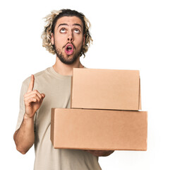 Young man with cardboard boxes in studio pointing upside with opened mouth.